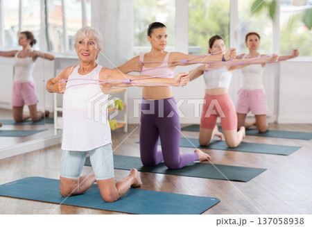 Group of women of different ages doing Pilates - kneeling and stretching their arms using an expander in gym Group of women of different ages doing Pilates - kneeling and stretching their arms using an expander in gym 137058938