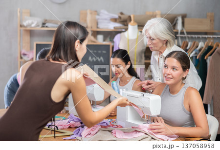 Girl stitching at machine and talking to groupmate in sewing workshop 137058958