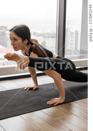 A woman in black activewear demonstrates a challenging yoga pose on a mat in front of a large window with a city view 137063244