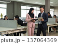 Modern office scene with diversity professionals collaborating. Woman in pink hijab suit taking notes while man in dark suit holding digital tablet. Colleagues work at office desks in open workspace 137064369