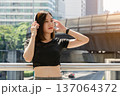 Young Asian woman with brown hair and dressed in athletic wear enjoying music through white headphones while standing on urban bridge or walkway 137064372