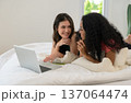 Two young women lie on bed enjoying time together while smiling and chatting in front of laptop in bedroom at home 137064474
