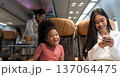 Black girl laughing joy fully on airplane while woman beside her smile at smart phone. Female flight attendant in background serves passengers. Airline transportation industry concept 137064475