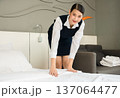 Smiling hotel Asian maid in dark blue uniform making bed with white linens in modern guest room 137064477