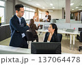 Professionals collaborate in modern office environment. Asian man in glasses and woman discussing at desk while colleagues working in background 137064478