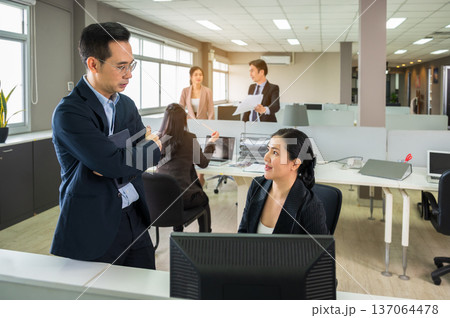 Professionals collaborate in modern office environment. Asian man in glasses and woman discussing at desk while colleagues working in background Professionals collaborate in modern office environment. Asian man in glasses and woman discussing at desk while colleagues working in background 137064478