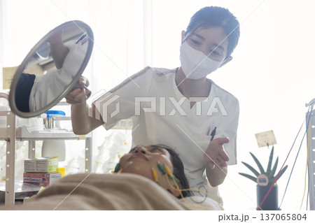 美容鍼に鍼電極低周波治療器をした状態を患者に見せて説明をする鍼灸師女性 美容鍼に鍼電極低周波治療器をした状態を患者に見せて説明をする鍼灸師女性 137065804