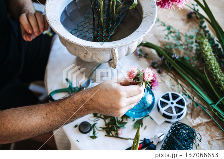 Close up of male hands holding pink aster flower over table with floral tools and white metal urn. slow flower movement, local flower farming, seasonal cut flowers, domestic flower industry Close up of male hands holding pink aster flower over table with floral tools and white metal urn. slow flower movement, local flower farming, seasonal cut flowers, domestic flower industry 137066653