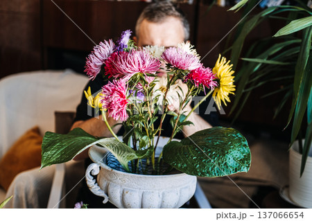 Man arranges bright aster flowers in ornate white pedestal urn. Sustainable mechanics, green living, garden-to-vase, environmentally conscious hobby. 137066654