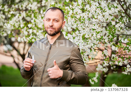 Man allergic using medical eyes drops, suffering from seasonal allergy at spring in blossoming garden. Portrait of handsome man showing thumbs up in front of blooming tree. Spring allergy concept. 137067328