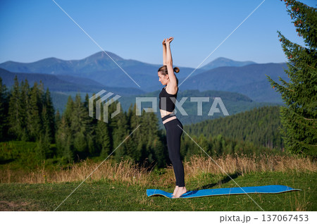 Woman practicing yoga outdoors in the mountains in a serene, natural setting. Female performing yoga pose on mat, with backdrop of beautiful mountain landscape at sunrise or sunset. Woman practicing yoga outdoors in the mountains in a serene, natural setting. Female performing yoga pose on mat, with backdrop of beautiful mountain landscape at sunrise or sunset. 137067453
