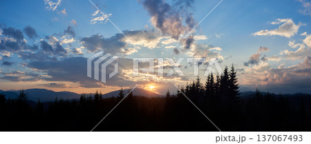 Sun sets behind range of distant mountains, casting golden rays through clouds. Sky, painted with shades of blue, pink, and gold, in tranquil evening. Silhouetted pine trees add depth to landscape. 137067493