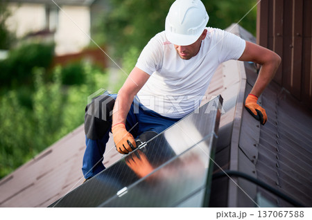 Worker building photovoltaic solar panel system on rooftop of house. Man engineer in helmets and gloves installing solar module with help of hex key outdoors. Renewable energy generation concept. Worker building photovoltaic solar panel system on rooftop of house. Man engineer in helmets and gloves installing solar module with help of hex key outdoors. Renewable energy generation concept. 137067588