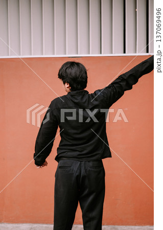 Person in Black Casual Attire with Arm Raised Against Orange Wall 137069496