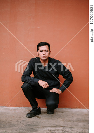 Serious young man in black squatting against a terracotta wall Serious young man in black squatting against a terracotta wall 137069685