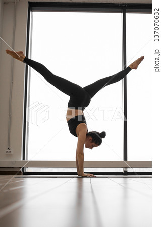 A woman in black athletic wear executes a challenging yoga pose, balancing on her hands with legs extended upwards against a bright window backdrop A woman in black athletic wear executes a challenging yoga pose, balancing on her hands with legs extended upwards against a bright window backdrop 137070632