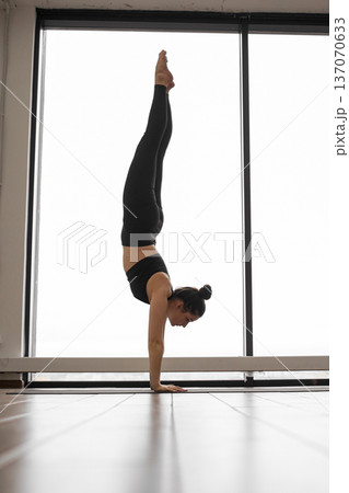 A woman in black athletic wear balances on her hands in a yoga studio with a large window behind her A woman in black athletic wear balances on her hands in a yoga studio with a large window behind her 137070633