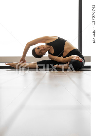 A woman in black activewear performs a side bend stretch on a yoga mat, showcasing flexibility and balance 137070743