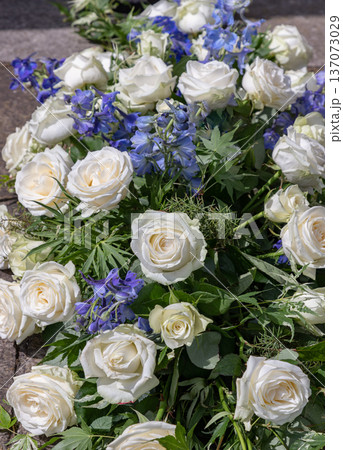 Full-bodied white roses and spires of cornflower delphinium are elegantly interwoven against a textured backdrop of fern and maple foliage, creating a luminous display of creamy petals and vibrant 137073029