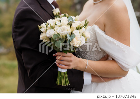 Elegant wedding couple embracing outdoors with white rose bouquet and lace dress in natural daylight 137073848