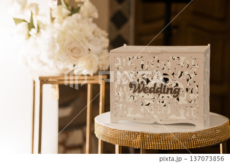 Elegant White Wedding Card Box with Ornate Laser Cut Design Placed on Gold-Rimmed Marble Table Near Floral Arrangement 137073856