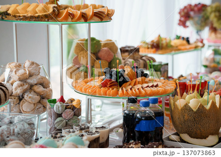 Vibrant Fruit and Dessert Buffet Display with Citrus Slices, Grapes, Candies, and Melon Centerpiece on Tiered Glass Stands Vibrant Fruit and Dessert Buffet Display with Citrus Slices, Grapes, Candies, and Melon Centerpiece on Tiered Glass Stands 137073863