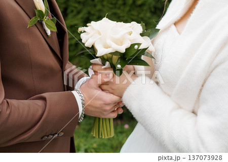 Close-up of wedding couple holding white calla lily bouquet with brown suit and white coat in garden setting Close-up of wedding couple holding white calla lily bouquet with brown suit and white coat in garden setting 137073928