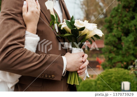 Elegant Wedding Couple Embracing Outdoors with White Calla Lilies and Brown Suit in Natural Garden Setting Elegant Wedding Couple Embracing Outdoors with White Calla Lilies and Brown Suit in Natural Garden Setting 137073929