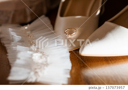 Elegant Wedding Ring Resting on White Bridal Shoes with Lace Garter on Wooden Surface Elegant Wedding Ring Resting on White Bridal Shoes with Lace Garter on Wooden Surface 137073957