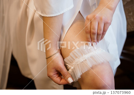Bride wearing white lace garter during wedding preparations, close-up of hands adjusting tulle accessory on leg 137073960
