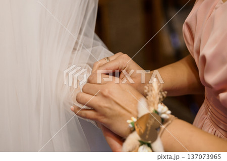 Close-up of a bridesmaid adjusting a bride's veil during wedding ceremony with soft pink and white tones and delicate floral wrist corsage 137073965