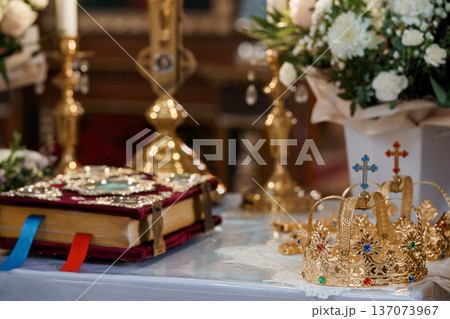 Golden Wedding Crowns and Ornate Religious Book on Altar with White Flowers and Candlelight in Church Interior Golden Wedding Crowns and Ornate Religious Book on Altar with White Flowers and Candlelight in Church Interior 137073967