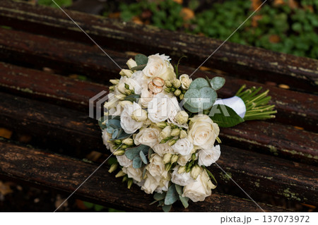 White Rose Wedding Bouquet with Gold Rings on Rustic Wooden Bench in Natural Garden Setting 137073972