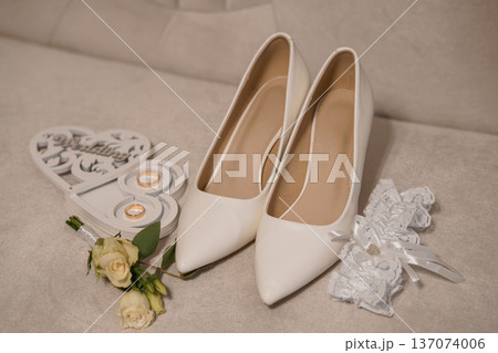 White Wedding Shoes with Lace Garter, Rings, Rose Bouquet, and Heart-Shaped Ornament on Beige Couch White Wedding Shoes with Lace Garter, Rings, Rose Bouquet, and Heart-Shaped Ornament on Beige Couch 137074006