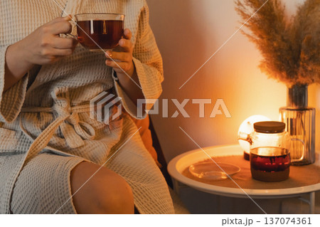 Woman in bathrobe holding a cup of tea, unwinding in a warm, comfortable home environment cozy relaxing for an evening or morning Woman in bathrobe holding a cup of tea, unwinding in a warm, comfortable home environment cozy relaxing for an evening or morning 137074361