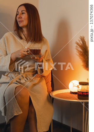 Woman in bathrobe enjoying a quiet moment, drinking a warm cup of tea in cozy home atmosphere with soft light Woman in bathrobe enjoying a quiet moment, drinking a warm cup of tea in cozy home atmosphere with soft light 137074366
