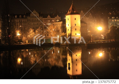 Illuminated historic tower rises beside a calm river at night, reflecting on still water 137075772