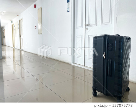 Black suitcase positioned in a well-lit hotel corridor with white walls, numbered door visible, and polished tile floor reflecting light Black suitcase positioned in a well-lit hotel corridor with white walls, numbered door visible, and polished tile floor reflecting light 137075778