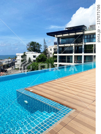 Rooftop swimming pool with clear blue water and wooden deck overlooking residential buildings and ocean under a bright blue sky in a coastal city 137075786