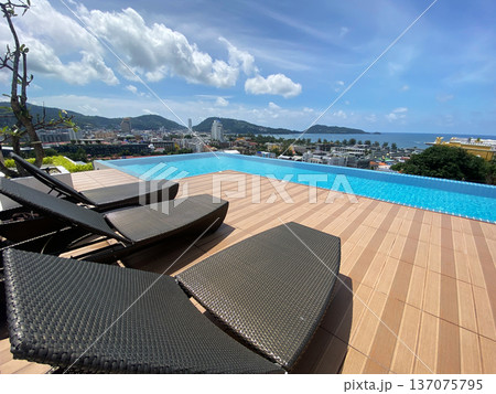 Luxury outdoor poolside view with modern lounge chairs overlooking the ocean and cityscape, surrounded by lush greenery and mountains in the background Luxury outdoor poolside view with modern lounge chairs overlooking the ocean and cityscape, surrounded by lush greenery and mountains in the background 137075795