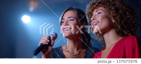 Two young women singing together on stage with microphone under bright spotlight in a joyful performance setting 137075976