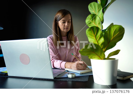 Woman writing in notebook while sitting at desk with laptop in home workspace 137076176