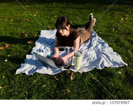Young woman working on laptop outdoors in park, remote freelance lifestyle, female typing on computer while lying on blanket, digital nomad productivity concept Young woman working on laptop outdoors in park, remote freelance lifestyle, female typing on computer while lying on blanket, digital nomad productivity concept 137078967