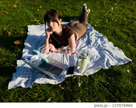 Young woman working remotely on laptop in sunny park, freelance digital nomad lifestyle, female typing on computer outdoors on blanket in green grass Young woman working remotely on laptop in sunny park, freelance digital nomad lifestyle, female typing on computer outdoors on blanket in green grass 137078968