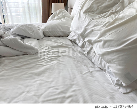 Unmade wrinkled white bed linen with crumpled pillows on a bed, natural light filtering through sheer curtains in a cozy bedroom setting Unmade wrinkled white bed linen with crumpled pillows on a bed, natural light filtering through sheer curtains in a cozy bedroom setting 137079464