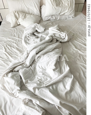 Unmade wrinkled white bed linen with crumpled sheets and pillows on a bed, showcasing a casual and relaxed bedroom setting 137079481