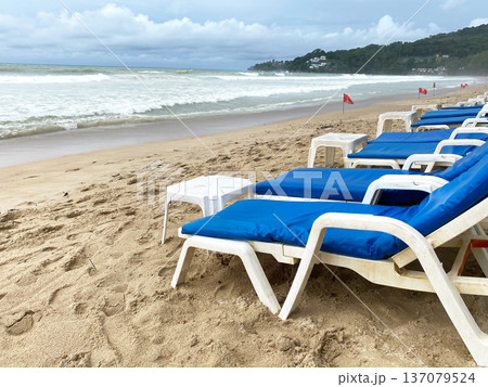 Beach scene featuring white lounge chairs with blue cushions arranged on sandy shore near ocean waves and distant green hills under a cloudy sky 137079524