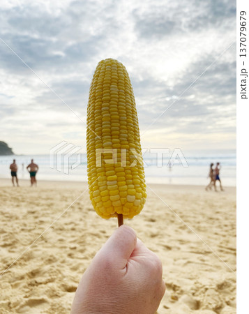 Hand holding a boiled corn on a stick against a beach backdrop with people enjoying the ocean waves and cloudy sky in the background Hand holding a boiled corn on a stick against a beach backdrop with people enjoying the ocean waves and cloudy sky in the background 137079679