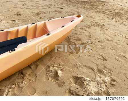 Orange kayak resting on sandy beach with visible footprints and textured sand, showcasing a tranquil outdoor setting ideal for water sports and recreation 137079692