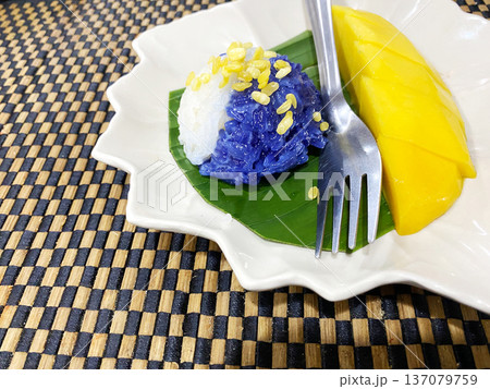 Thai mango sticky rice dessert featuring blue and white glutinous rice topped with mung beans, served with fresh mango slices on a decorative plate with a fork Thai mango sticky rice dessert featuring blue and white glutinous rice topped with mung beans, served with fresh mango slices on a decorative plate with a fork 137079759